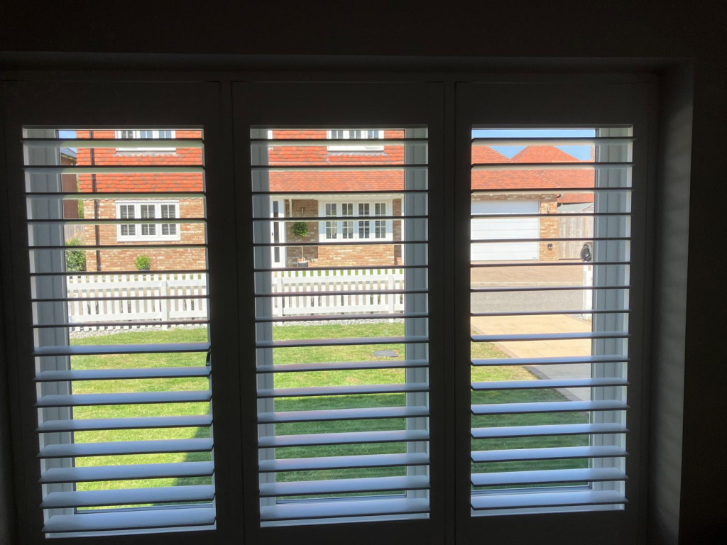 View through white shuttered windows with a stylish home décor solution: cassette roller blinds framing a sunny lawn and red-roofed brick houses.