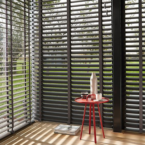 A small red table with a vase and cups sits by large windows with made to measure Venetian blinds, letting in sunlight and revealing greenery.