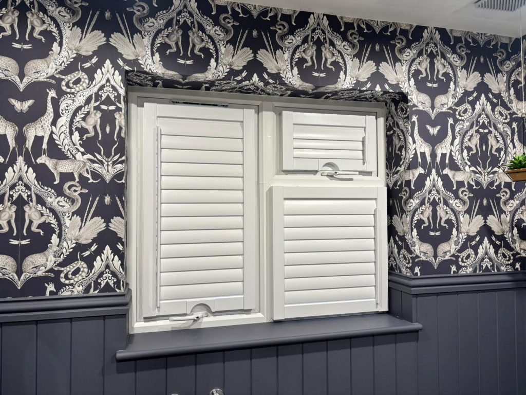 White plantation shutters by JLS Blinds on a window with ornate black and white wallpaper featuring birds and floral patterns.