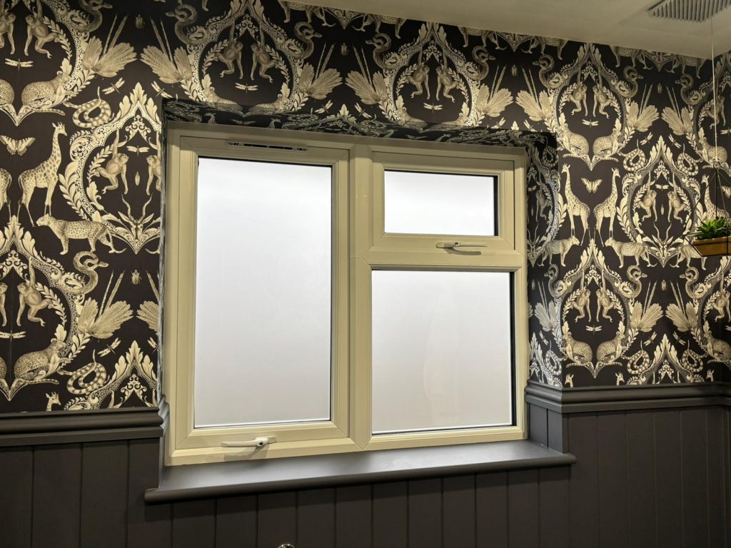 A white-framed window with frosted glass features JLS blinds, set amid ornate black and white wallpaper and dark panelled walls.