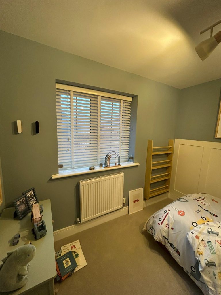 A neatly organised children’s bedroom with a window fitted with JLS blinds, a radiator, small bed, shelf, and toys on a low table.