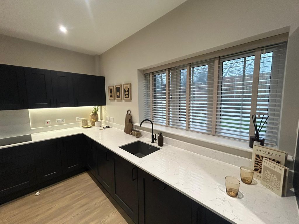 Modern kitchen with black cupboards, white marble worktops, large window with JLS blinds, and minimalist decor on the worktops and walls.