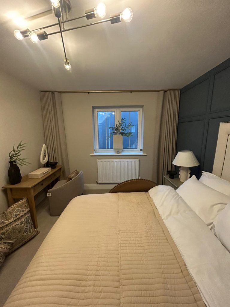 Modern bedroom with beige bedding, a desk by the window featuring JLS blinds, and decorative vases with greenery.