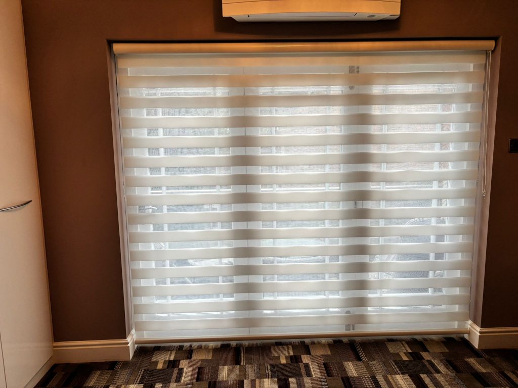 Large window with modern JLS blinds, horizontal stripes partially closed, letting in diffused light; patterned carpet beneath.