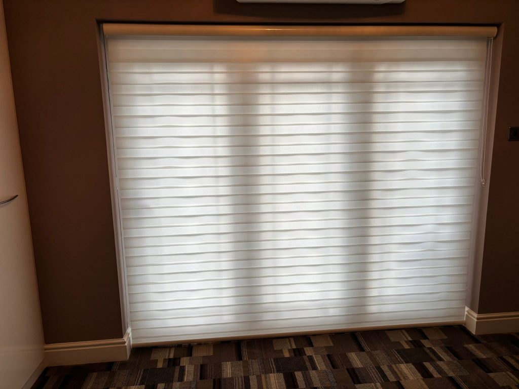 JLS Blinds' white fabric blinds cover a large window, diffusing light into a room with patterned carpet and beige walls.