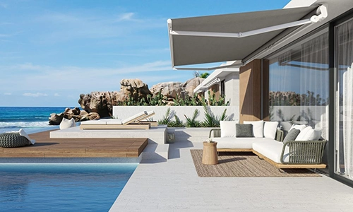 Modern beachfront patio with a pool, white sun loungers, a sofa set on a rug, wooden decking, and retractable awning, overlooking blue sea and rocky shoreline under a clear sky.