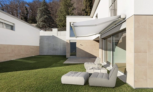 Modern house with beige and white exterior, featuring large glass doors, a retractable awning, and a grassy garden with outdoor lounge chairs and a small table. Trees and additional buildings are in the background.