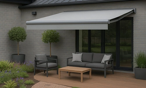 A modern patio with grey outdoor furniture, including a sofa, armchair, and wooden coffee table, sits under a large grey retractable awning. Two potted trees and plants decorate the area near glass doors.