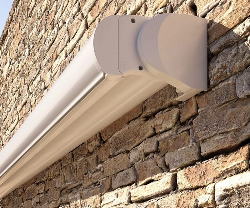 A retractable awning with a closed casing is mounted on a textured stone wall, shown from an angled perspective. This streamlined design blends with the outdoor setting—perfect for enhancing spaces with Conservatory Awnings.