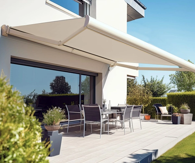 A modern patio with potted plants, outdoor dining furniture, and a large beige retractable conservatory awning attached to a white house, providing shade on a sunny day.