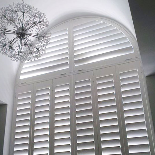 White plantation shutters, made to measure for a large arched window, let sunlight filter through the slats. A modern, spherical chandelier with radiating metal rods hangs in the upper left corner.