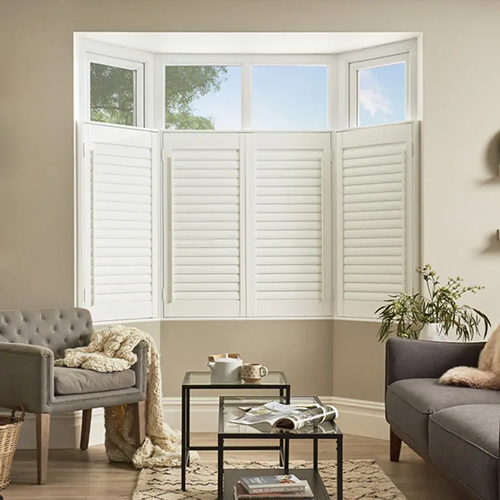 A cosy living room with a bay window fitted with Made to Measure Shutters, a grey armchair draped with a blanket, a grey sofa, a glass coffee table, and a potted plant on the side table.