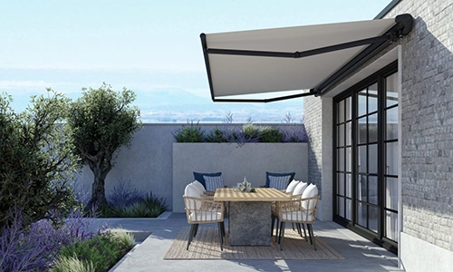A modern patio with a wooden dining table and four cushioned chairs sits next to a brick house. A large commercial awning provides shade, whilst plants and shrubs surround the inviting area.