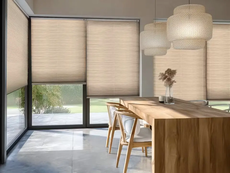 A modern dining area with large windows covered by beige cellular & pleated blinds, a wooden table with four chairs, two decorative pendant lights, and a vase with dried flowers on the table. Natural light fills the space.