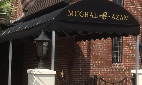 A black commercial awning with Mughal e Azam written in gold letters covers the entrance of a brick building, next to a vintage-style black lamp post.