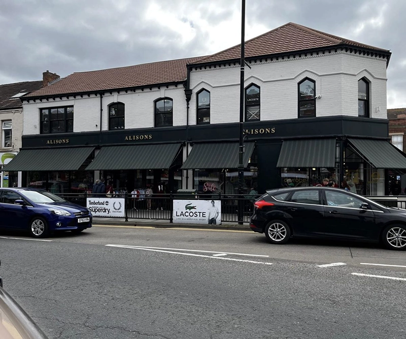 A two-storey white building houses ALISONS, a shop with dark green commercial awnings over its windows. Several cars are parked in front, and a cloudy sky is visible above.