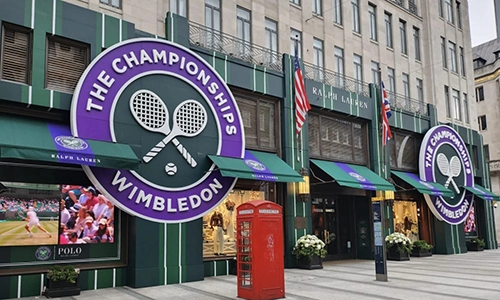 A building decorated with large Wimbledon Championship signs, commercial awnings in green, and flags. A Ralph Lauren shop is visible, with a red telephone box and tennis imagery featured in the window displays.