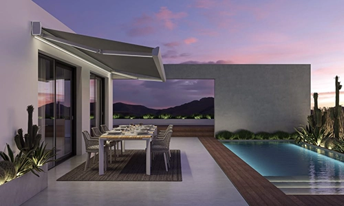 Modern patio with a dining table set for eight under a retractable awning, beside a pool, with desert plants and mountains visible at dusk.