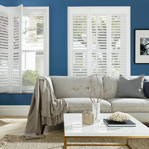 A modern living room with a light grey sofa, cosy blanket and cushions, a white coffee table with decor, and large windows fitted with Made to Measure Shutters letting in natural light against a blue wall.