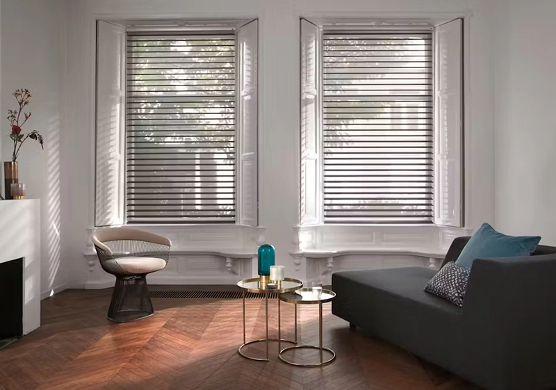 A bright, modern living room features two large windows with Silhouette Shades, a grey sofa with cushions, a round side chair, nested gold tables with vases, and wooden herringbone flooring. Sunlight streams softly through the shades.