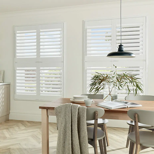 A bright dining area with a wooden table, four grey chairs, an open book, a plant centrepiece, and a hanging black lamp. Large windows with Made to Measure Shutters let in natural light. A grey blanket is draped over a chair.