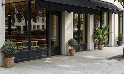 Street view of a modern café with large glass windows, sleek commercial awnings, and potted plants by the entrance. Wooden tables and chairs are visible inside through the windows.