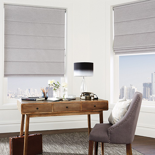 A home office scene with a wooden desk, grey upholstered chair, black table lamp, and two large windows fitted with made-to-measure Roman blinds overlooking a city skyline. A briefcase sits on the floor beside the desk.