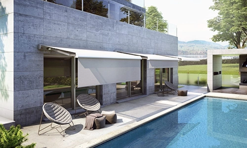 Modern house with large windows and two commercial awnings above patio doors, next to a swimming pool. The patio offers two round chairs and a small table, with trees, grass, and distant hills providing a scenic backdrop.