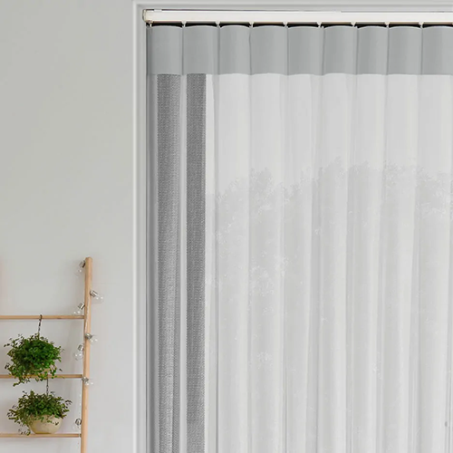 A sheer white curtain with a solid grey border hangs in front of a window, whilst made to measure vertical blinds offer privacy. On the left, a wooden ladder-style shelf holds potted green plants against a light-coloured wall.