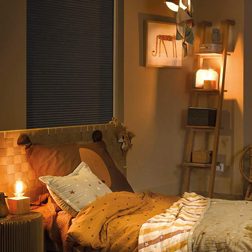 A cosy children's bedroom with warm lighting, a bed with star-patterned bedding, a teddy bear-themed headboard, a small lit lamp, a wooden ladder shelf with decor, and a framed giraffe picture on the wall.