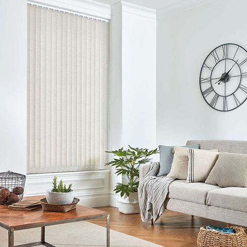A modern living room with a large made-to-measure vertical blind, a grey sofa with neutral cushions, a potted plant, a wall clock with Roman numerals, and a wooden coffee table with a basket and potted plants.