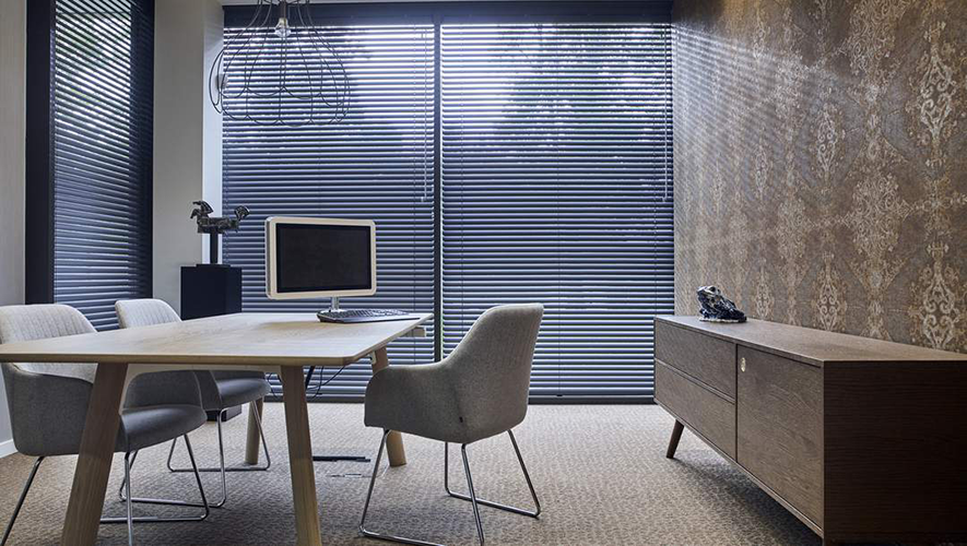Modern office with a wooden desk, two grey chairs, and a small table with a monitor. Floor-to-ceiling windows are covered by commercial blinds, letting in filtered daylight. A sideboard sits against the right wall.