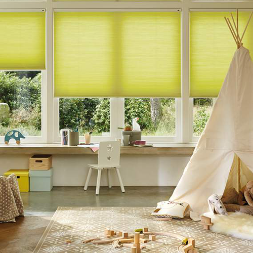 A bright playroom with large windows covered by yellow made to measure roller blinds, a white desk and chair, a toy tipi, scattered wooden toys, and a patterned rug on the floor. Greenery is visible outside the windows.