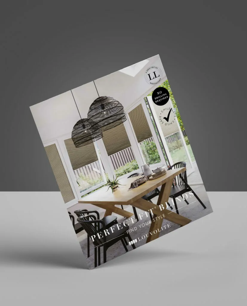 A magazine stands upright, featuring a modern dining room with large windows, woven pendant lights, a wooden table, black chairs, and beige window blinds. The cover text reads PERFECT BLINDS TO FIND YOUR STYLE by Louvolite.