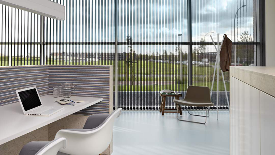 Modern office space with a white desk, chairs, a laptop, and spectacles. Large windows with commercial blinds overlook a grassy area. A grey lounge chair and coat stand are in the corner. Natural light fills the room.