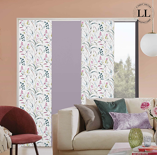 A modern living room with a beige sofa and colourful cushions, a mauve chair, and large windows covered with Made to Measure Panel Blinds Kent in floral and lavender designs. A round white pendant light hangs from the ceiling.
