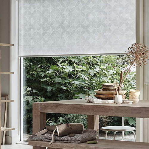 A dining area with a wooden table and bench, set with plates and decorative items, in front of a large window partially covered by a patterned roller blind, overlooking lush green plants outside.