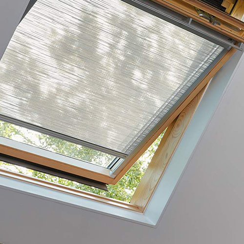 A skylight window set in a sloping ceiling is partially open, with a semi-transparent white blind pulled down. Sunlight and leaves from nearby trees are visible through the glass.