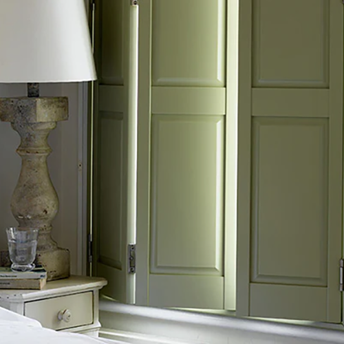 A close-up of a bedside table with a stone lamp, stacked books, and a glass of water in front of pale green Made to Measure Shutters, with light streaming through the edges.