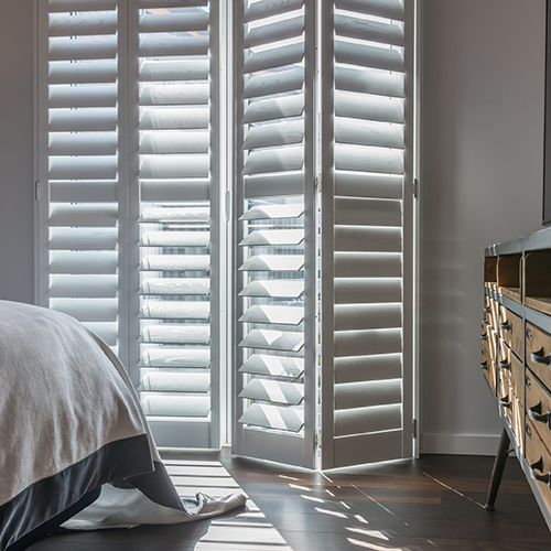 White, Made-to-Measure Shutters are partially open, letting sunlight stream into a modern bedroom with dark wooden floors. Sunlight creates patterns on the floor and bed, and a reflective chest of drawers is visible to the right.