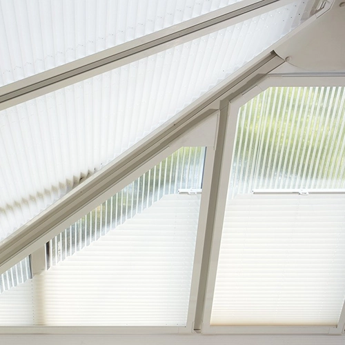 A close-up view of angled windows with Louvolite Perfect Fit International Blinds, their white slats partially open, allowing soft light and a glimpse of greenery outside to filter through the ribbed glass.
