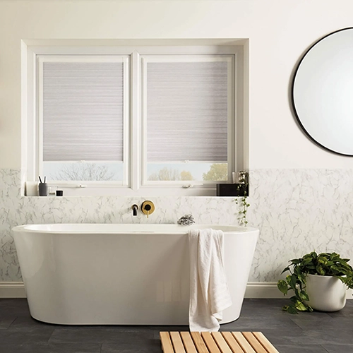 A modern bathroom with a white freestanding bath, a towel draped over the side, a wooden bath mat, a round mirror, two large windows with Perfect Fit Shutters Kent, and a potted plant on the floor.