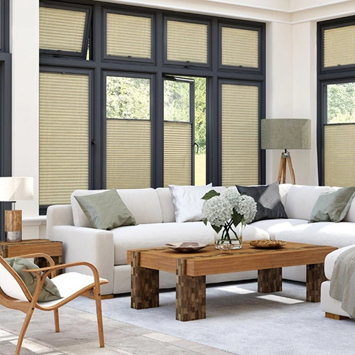 A bright living room with large windows dressed in Perfect Fit Pleated Blinds, a light-coloured sectional sofa with green and grey cushions, a wooden coffee table with white flowers, an armchair, and a floor lamp—all in neutral tones and natural light.