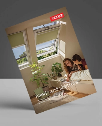 A brochure tilted against a grey background shows two people lying on a bed under roof windows with green blinds. The cover reads VELUX. Blinds and shutters and features indoor plants and sunlight streaming in.
