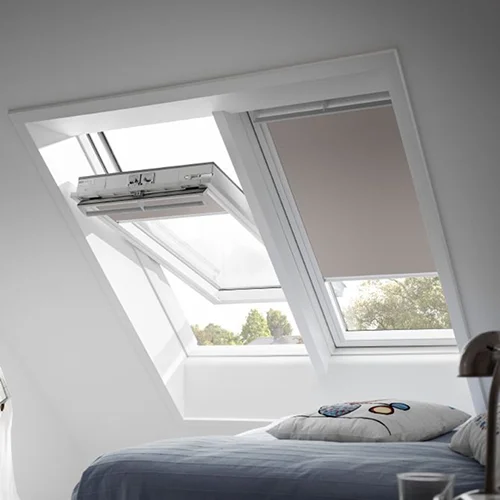 A modern loft bedroom with two slanted windows, one open and fitted with venetian blinds, above a bed with light blue bedding.