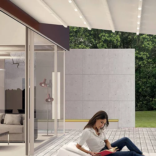A woman sits and reads on a patio with modern architecture, greenery, and sleek JLS blinds in the background.