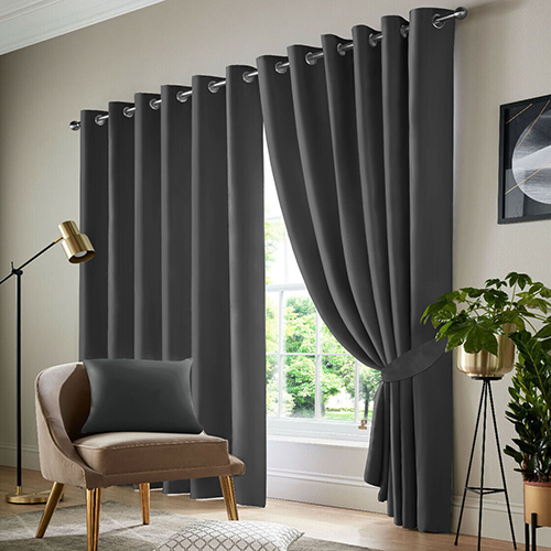 A modern living room with dark grey made-to-measure curtains, one side pulled back, covering a large window. The room features a beige armchair with a dark cushion, a gold floor lamp, a plant, and white walls with minimal decor.