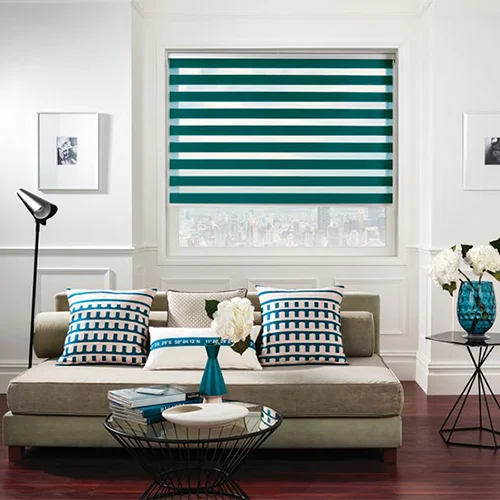 Modern living room with teal striped JLS blinds, patterned cushions, a beige sofa, and floral décor on glass and metal tables.