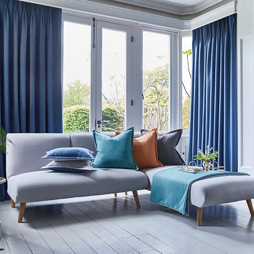 Modern living room with a grey sofa, blue curtains, teal accents, and JLS Blinds on large windows overlooking a garden.