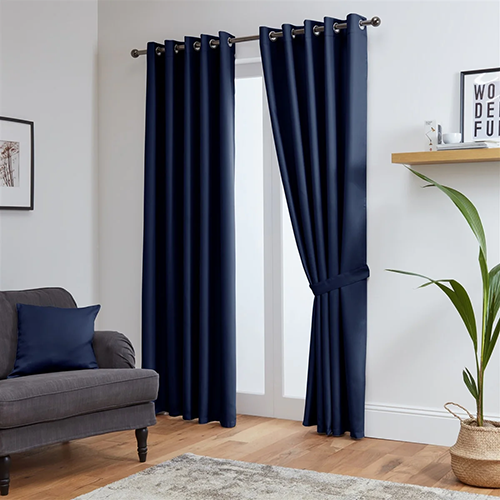 A modern living room with navy blue made-to-measure curtains on a window, one curtain tied back. There is a grey sofa with a blue cushion, a plant, a beige rug, and wooden floor. A wall shelf and framed art are visible.